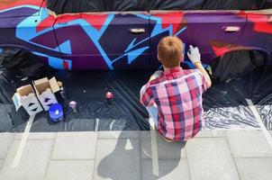 en ung rödhårig graffiti konstnär målarfärger en ny färgrik graffiti på de bil. Foto av de bearbeta av teckning en graffiti på en bil närbild. de begrepp av gata konst och olaglig vandalism