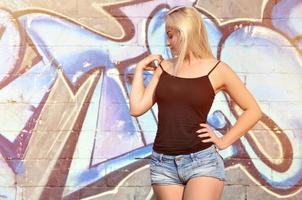 sexig caucasian blond flicka i denim shorts och svart tank topp Framställ mot graffiti vägg i de dagtid utomhus foto