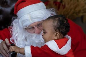 afrikansk amerikan bebis är öppning de jul närvarande med santa claus på natt för säsong firande foto