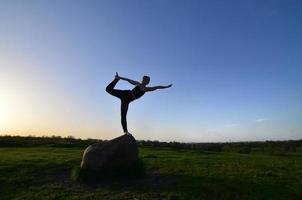 silhuett av ung blond flicka i sport kostym öva yoga på en pittoresk grön kulle i de kväll på solnedgång. de begrepp av utövar och friska livsstilar foto