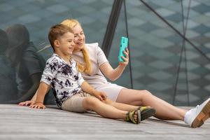 Lycklig blond kvinna och liten pojke Sammanträde på terrass och framställning selfie på smartphone. mor och son njut av tid tillsammans. positiv ung mamma utgifterna tid med henne söt barn. har roligt. familj. foto