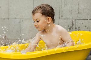 söt liten pojke badning i gul badkar utomhus. Lycklig barn är stänk, spelar med vatten och har roligt. sommar säsong och rekreation. vistas Häftigt i de sommar värme. vatten roligt i bakgård. foto