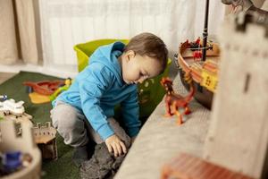 liten caucasian pojke med blå luvtröja spelar med färgrik leksaker på Hem. barn har roligt. Lycklig och glad unge spelar med fartyg, dinosaurier, slott. fritid aktivitet, inhemsk liv. mysigt rum. foto