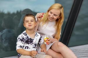 Lycklig blond kvinna och liten pojke Sammanträde på terrass och äter sötsaker. mor och son njut av tid tillsammans. positiv ung mamma spelar, utgifterna tid med henne söt barn, skrattande, har roligt. familj. foto