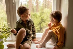 barn Sammanträde på fönsterkarm och väntar för någon kommer. två bröder, vänner. söt förskola barn ensam på Hem. Pojkar är väntar för deras mor eller far. ensamhet. upptagen föräldrar. foto