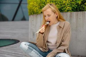 ung skön blond kvinna äter sushi utomhus, på de trä- terrass, förbi modern byggnad i de stad. gott mat till gå. flicka har lunch ha sönder, utgifterna tid utanför och äter asiatisk mat. stad liv foto