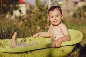 söt liten pojke badning i badkar utomhus i trädgård. Lycklig barn är stänk, spelar med vatten och har roligt. sommar säsong och rekreation. vistas Häftigt i de sommar värme. vatten roligt i bakgård. foto