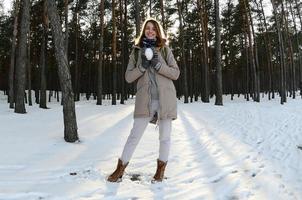 en ung och glad caucasian flicka i en brun täcka innehar en snöboll i en snötäckt skog i vinter. fisköga Foto