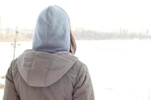 en ung caucasian flicka i en brun täcka stirrande in i de distans på de horisont linje mellan de himmel och de frysta sjö i vinter- foto