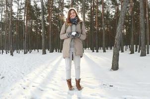 en ung och glad caucasian flicka i en brun täcka innehar en snöboll i en snötäckt skog i vinter. fisköga Foto