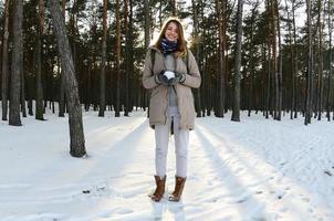 en ung och glad caucasian flicka i en brun täcka innehar en snöboll i en snötäckt skog i vinter. fisköga Foto