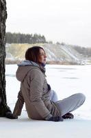 en ung caucasian flicka i en brun täcka stirrande in i de distans på de horisont linje mellan de himmel och de frysta sjö i vinter- foto