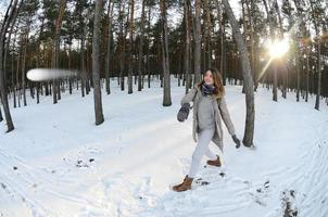 en ung och glad caucasian flicka i en brun täcka är kasta en snöboll i en snötäckt skog i vinter. spel med snö i de öppen luft. fisköga Foto