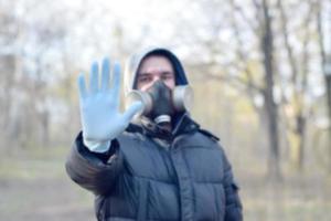 suddig porträtt av ung man i skyddande gas mask och sudd disponibel handskar visar sluta gest utomhus i vår trä foto