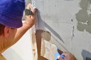 femtio år gammal manuell arbetstagare med vägg putsning verktyg renovera hus. murare renovera väggar och hörn med spatel och plåster. vägg mosa installation. konstruktion efterbehandling Arbetar foto