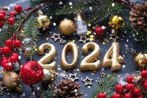 de gyllene siffror 2024 tillverkad av ljus på en svart sten skiffer bakgrund är dekorerad med en festlig dekor av stjärnor, paljetter, gran grenar, bollar och girlanger. hälsning kort, Lycklig ny år. foto