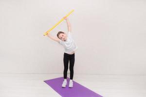 söt liten flicka står på en gymnastiska matta innehav upp en gymnastiska pinne och lutar sig, barns kondition foto