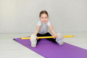 söt liten flicka Sammanträde på en matta och håller på med övningar med en gymnastiska pinne, barn kondition foto