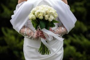de brud i en vit bröllop klänning är innehav en bukett av vit blommor - pioner, rosor. bröllop. brud och brudgum. delikat Välkommen bukett. skön dekoration av bröllop med löv foto