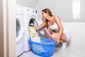 ung kvinna tar tvätt ut av tvättning maskin på Hem. kvinna läser in förberedelse tvättning maskin i badrum kläder i de tvättning maskin foto