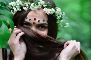 porträtt av ett emotionell ung flicka med en blommig krans på henne huvud och skinande ornament på henne panna. söt brunett Framställ i en spirande skön skog i de dagtid på en bra dag foto