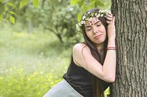 porträtt av ett emotionell ung flicka med en blommig krans på henne huvud och skinande ornament på henne panna. söt brunett Framställ i en spirande skön skog i de dagtid på en bra dag foto