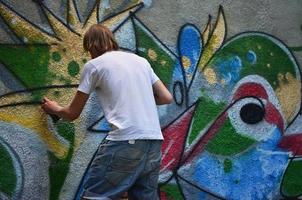 Foto av en ung kille i denim shorts och en vit skjorta. de kille drar på de graffiti vägg en teckning med aerosol målarfärger av olika färger. de begrepp av huliganism och skada till fast egendom