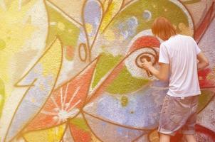 Foto av en ung kille i denim shorts och en vit skjorta. de kille drar på de graffiti vägg en teckning med aerosol målarfärger av olika färger. de begrepp av huliganism och skada till fast egendom