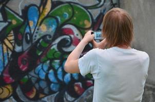 Foto i de bearbeta av teckning graffiti på ett gammal betong vägg. en ung, långhårig blond kille tar bilder av hans avslutad teckning på en smartphone. gata konst och vandalism begrepp