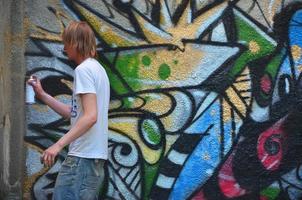 Foto av en ung kille i denim shorts och en vit skjorta. de kille drar på de graffiti vägg en teckning med aerosol målarfärger av olika färger. de begrepp av huliganism och skada till fast egendom