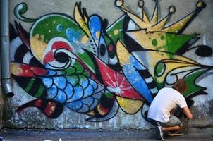 Foto av en ung kille i denim shorts och en vit skjorta. de kille drar på de graffiti vägg en teckning med aerosol målarfärger av olika färger. de begrepp av huliganism och skada till fast egendom