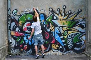 Foto av en ung kille i denim shorts och en vit skjorta. de kille drar på de graffiti vägg en teckning med aerosol målarfärger av olika färger. de begrepp av huliganism och skada till fast egendom