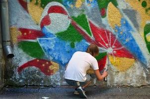 Foto av en ung kille i denim shorts och en vit skjorta. de kille drar på de graffiti vägg en teckning med aerosol målarfärger av olika färger. de begrepp av huliganism och skada till fast egendom