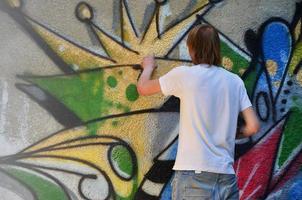Foto av en ung kille i denim shorts och en vit skjorta. de kille drar på de graffiti vägg en teckning med aerosol målarfärger av olika färger. de begrepp av huliganism och skada till fast egendom