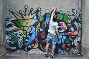 Foto av en ung kille i denim shorts och en vit skjorta. de kille drar på de graffiti vägg en teckning med aerosol målarfärger av olika färger. de begrepp av huliganism och skada till fast egendom