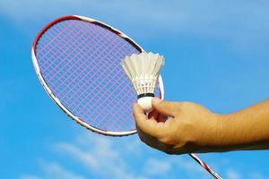 en person innehav en fjäderboll i främre av en badminton racket på de himmel bakgrund. begrepp för utomhus- badminton spelar i fri gånger, mjuk och selektiv fokus. foto