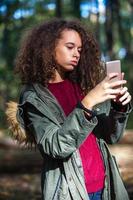 söt lockigt hår tonåring flicka med smartphone i höst skog foto