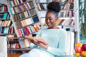 ung skön afrikansk amerikan flicka läsning en bok på de soffa med de bibliotek bokhyllor i de tillbaka. skön kvinna på en vit soffa läsning en bok foto