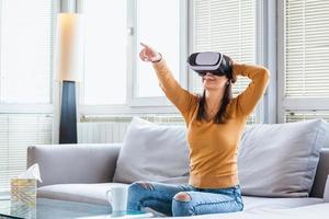 kvinna med virtuell verklighet glasögon. leende ung kvinna använder sig av vr headsetet på Hem på soffa. kvinna njuter virtuell verklighet på Hem. skön kvinna bär vr virtuell verklighet headsetet med gränssnitt. foto