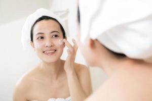 Lycklig ung asiatisk kvinna applicering ansikte lotioner medan bär en handduk och rörande henne ansikte i badrum foto