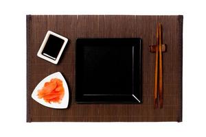 tömma svart fyrkant tallrik med ätpinnar för sushi, ingefära och soja sås på mörk bambu matta bakgrund. topp se med kopia Plats för du design foto