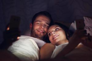 ung par använder sig av smartphones i säng på natt foto