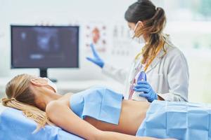 läkare i mask håller på med ultraljud buk testa till kvinna patient foto