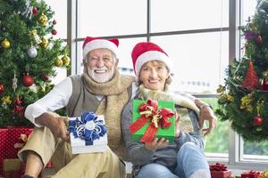 Lycklig leende senior caucasian par fira jul tillsammans medan utbyte deras närvarande i lycka och spänning på Hem med röd santa hatt och jul träd foto