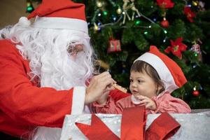 caucasian bebis är få närvarande från santa claus på natt förbi de fullt dekorerad jul träd för säsong firande begrepp foto