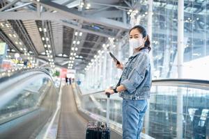 asiatisk kvinna turist bär ansikte mask och innehav smartphone på rulltrappa på flygplats terminal under coronavirus eller covid-19 utbrott. foto