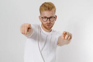 kille pekar på dig med fingrar isolerad på vit bakgrund. ung snygg rödhårig man, rött skägg och glasögon. vit t-shirt syns på dig. framgång och självförtroende koncept. kopieringsutrymme. selektiv fokusering foto