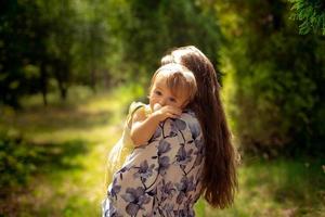 söt liten bebis flicka i yellos klänning kramar henne mor på de grön trädgård foto