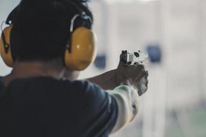 asiatisk skytten man bär ljud inställande hörlurar och svart kläder praktiserande skytte kort pistol på de skytte räckvidd. skytte sporter för meditation och självförsvar, rekreations aktiviteter. foto