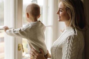 mor och henne liten bebis son bär värma tröjor ser på de fönster foto